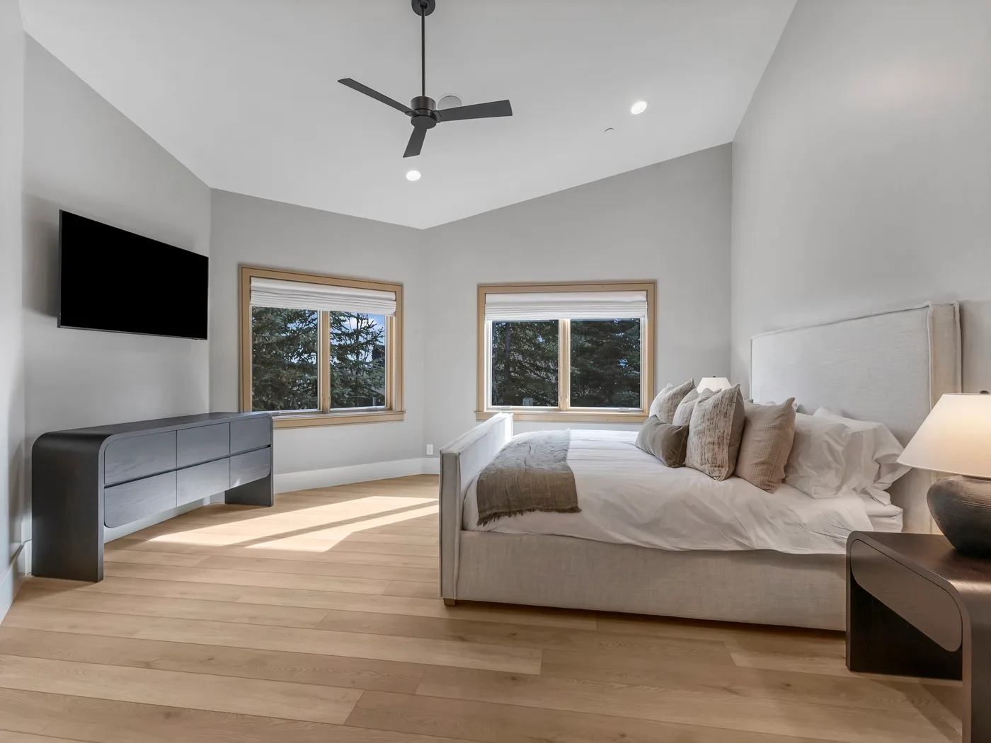 A bright bedroom with a bed, wall-mounted television, and two windows.
