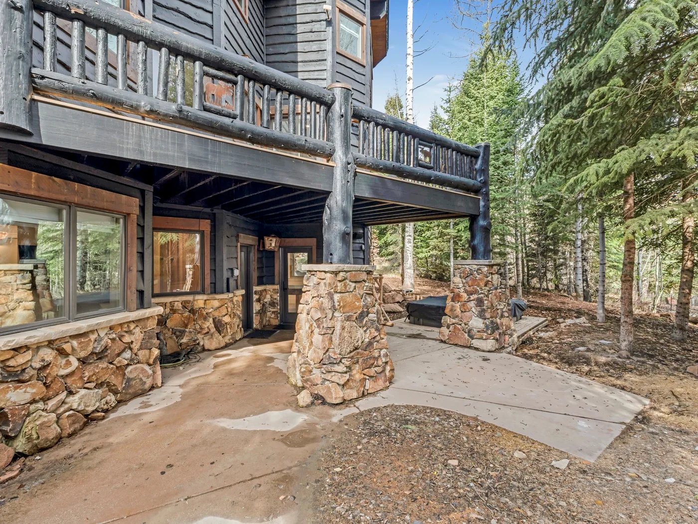 A stone-paved patio sits beneath a wooden balcony beside a dark-sided cabin.