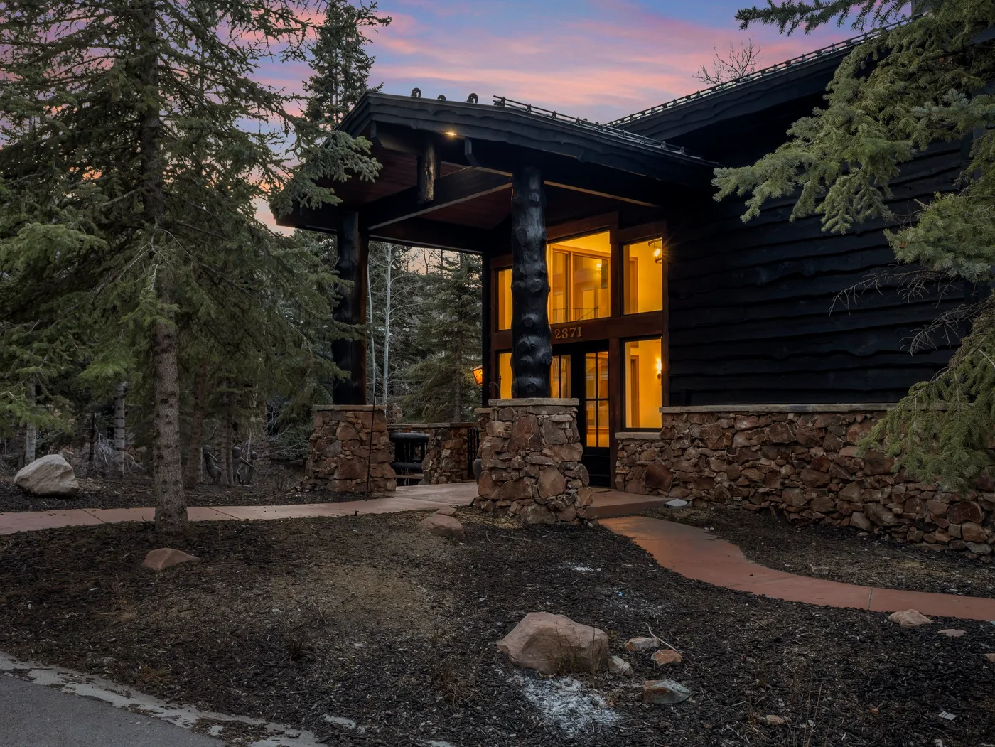 A dark timber lodge entrance glows warmly among tall evergreen trees at dusk.