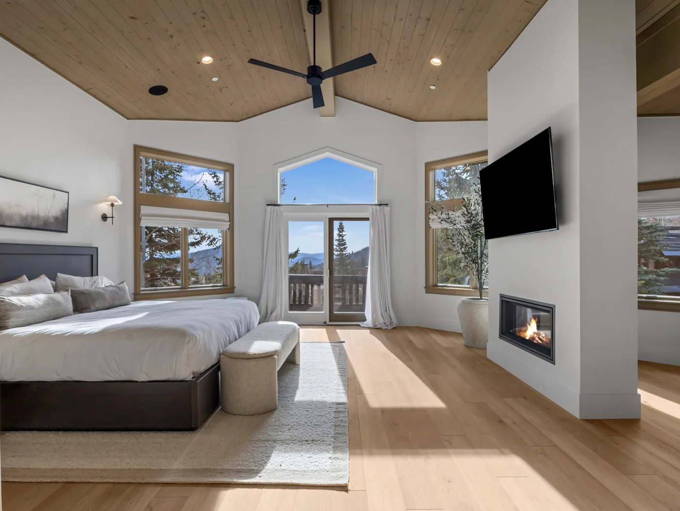 A bright bedroom with a large bed, wood flooring, and a fireplace beneath a wall-mounted television.