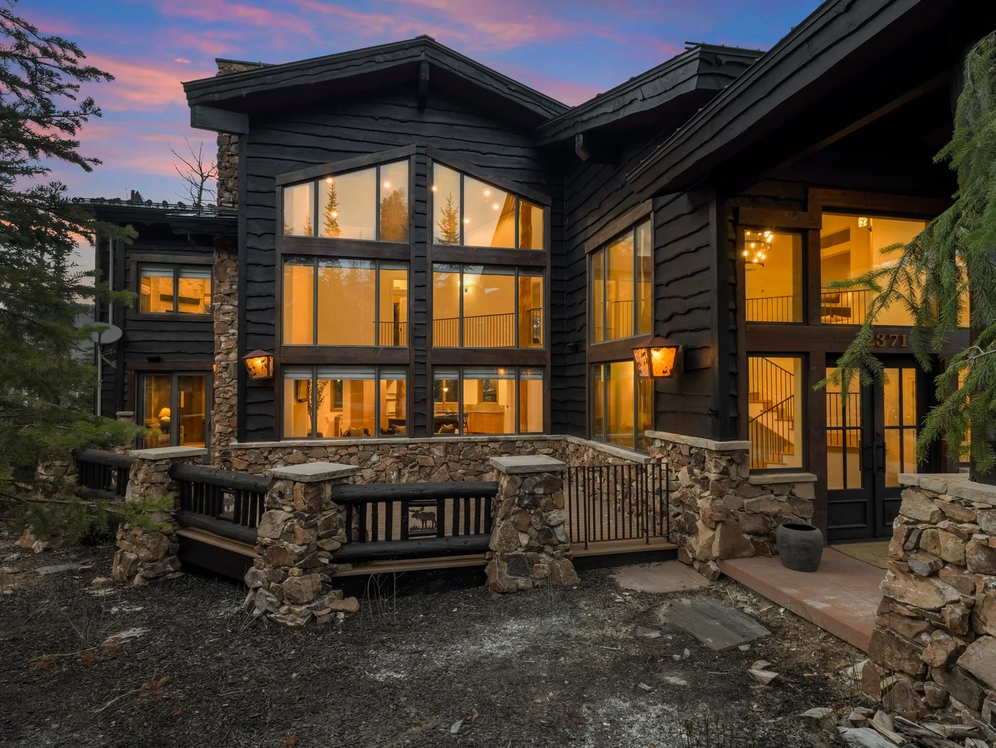 A large dark-clad house with illuminated windows and a stone terrace at dusk.