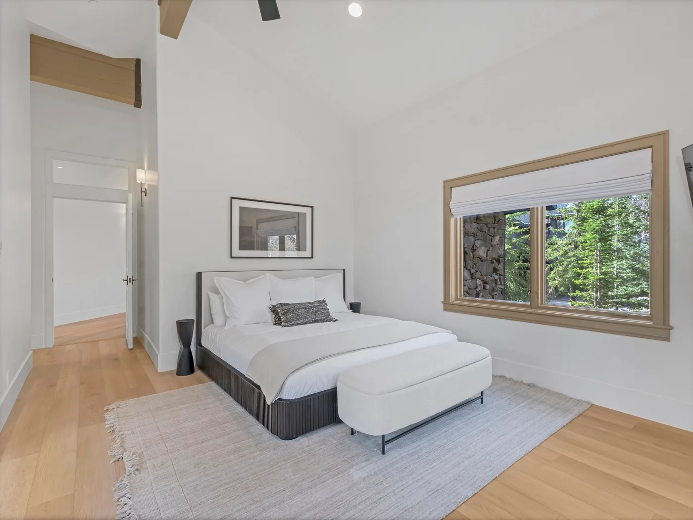 A bright bedroom with a bed, a large window, and light wood flooring.