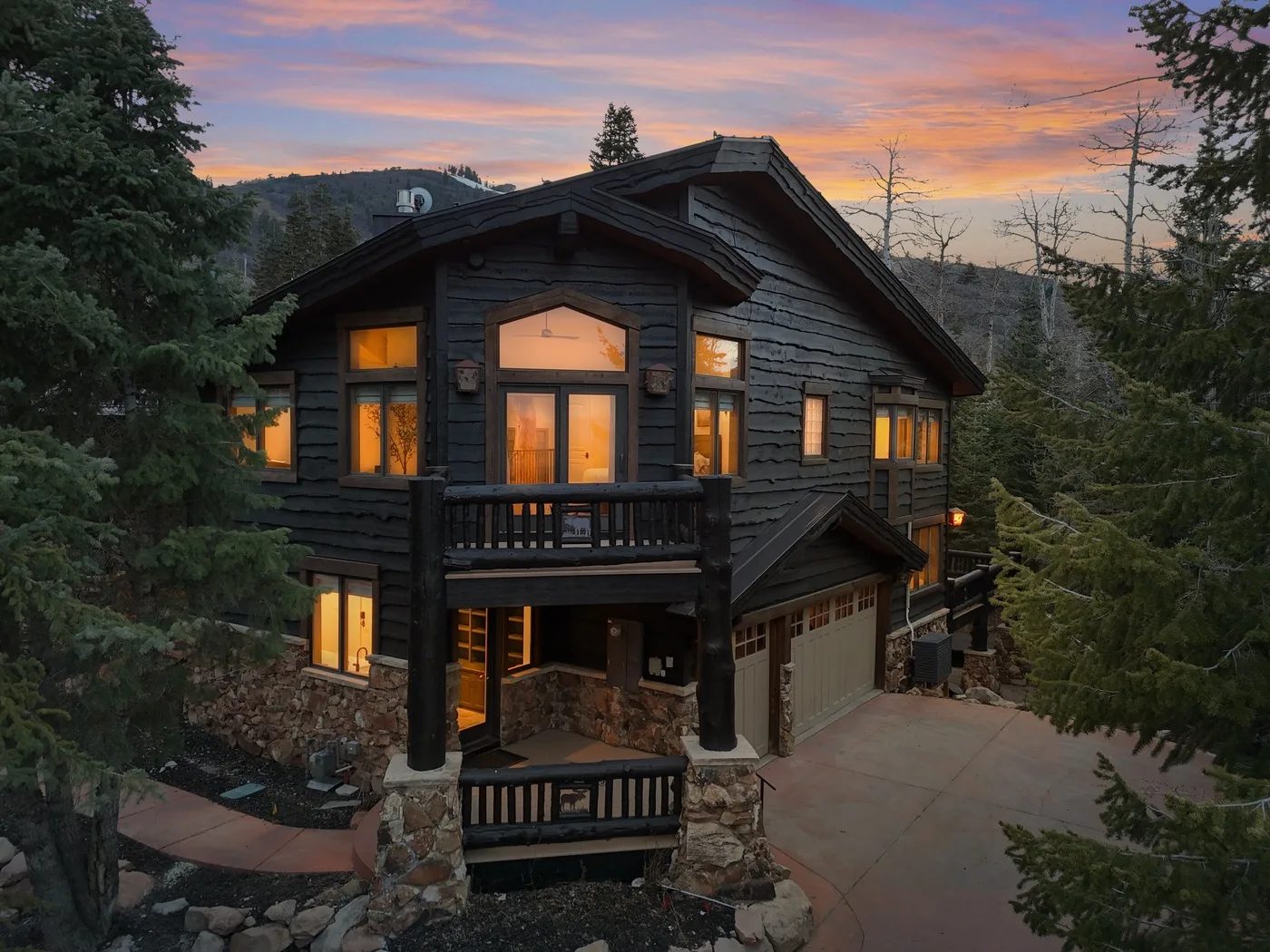 A dark timber house with a balcony and garage stands among trees at sunset.