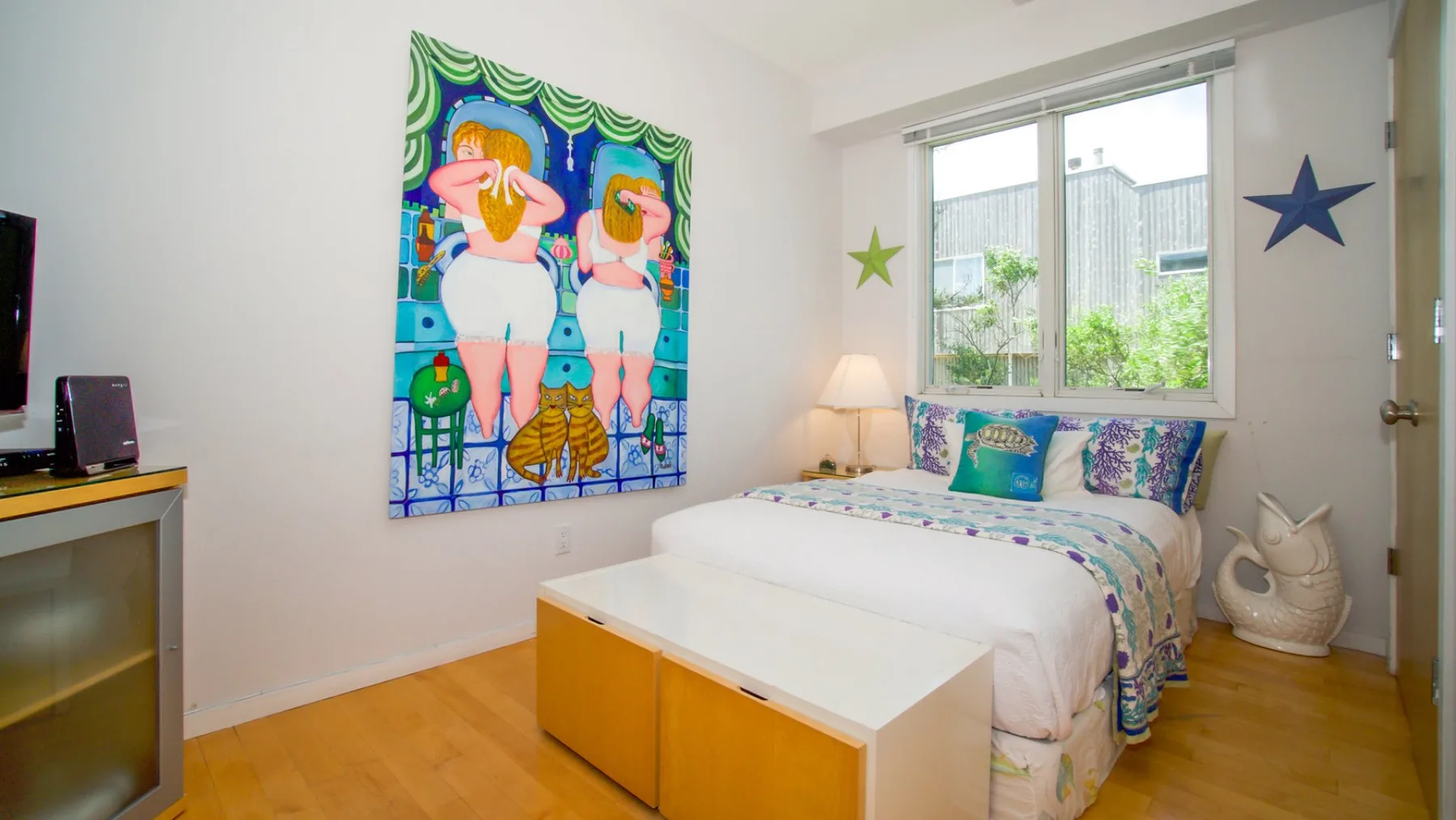 Un dormitorio luminoso con una cama bajo una ventana grande y arte colorido en la pared.