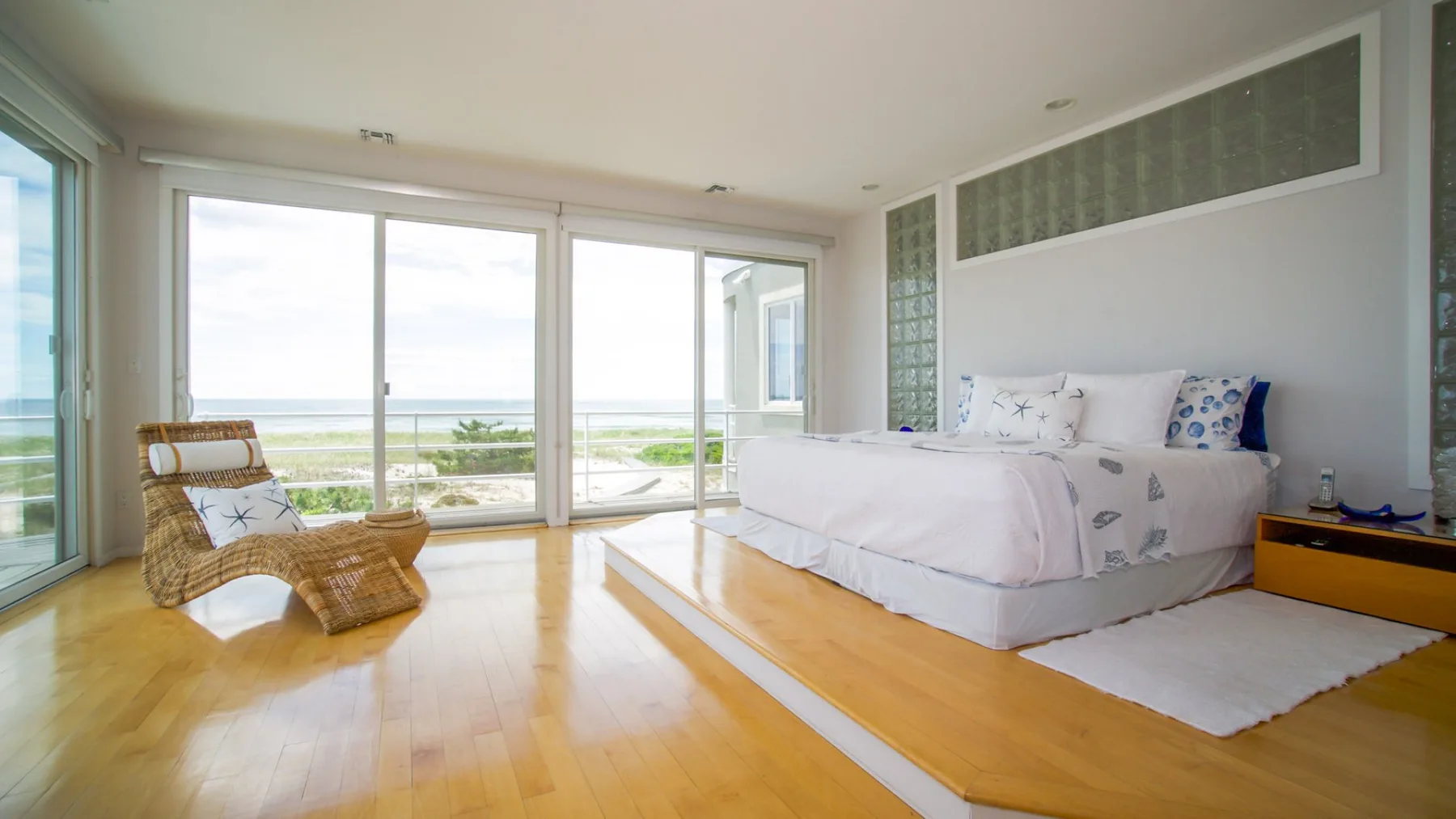 A bright bedroom with a bed, woven seating, and large windows overlooking the water.