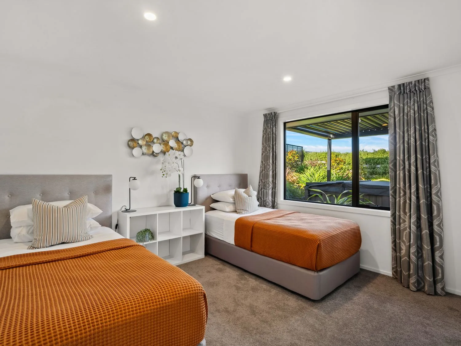 A bright bedroom with two beds, orange bedding, and a large window.