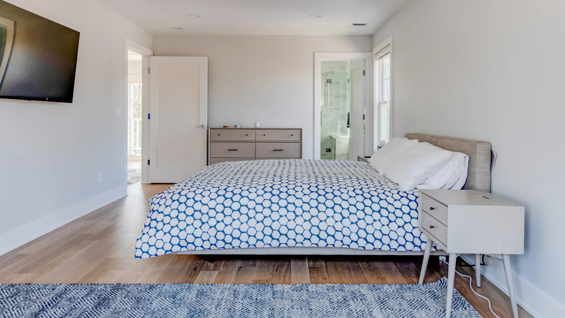 Un dormitorio luminoso con una cama, una cómoda y un televisor en la pared.