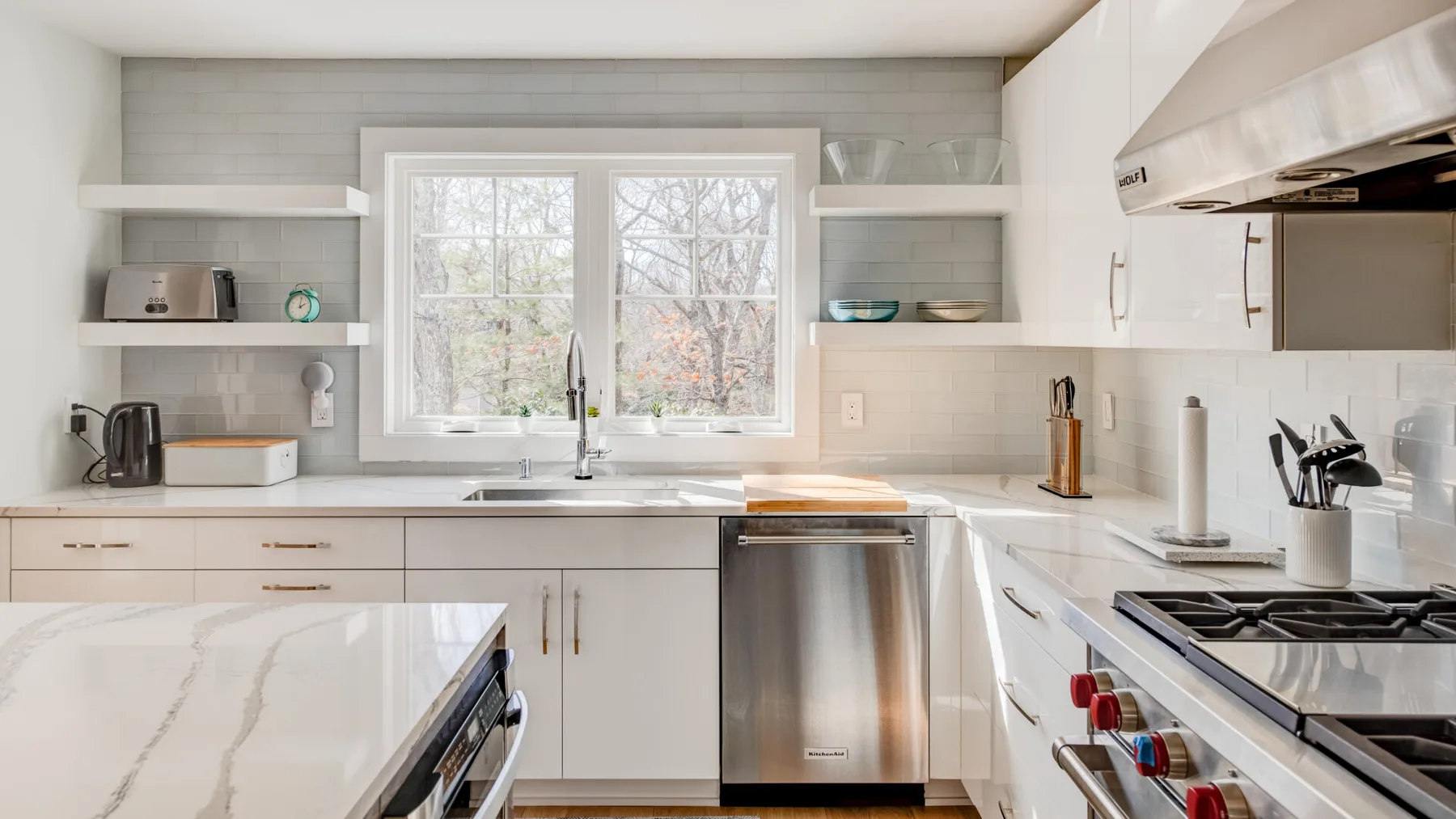 Una cocina luminosa con gabinetes blancos, una ventana central y electrodomésticos de acero inoxidable.