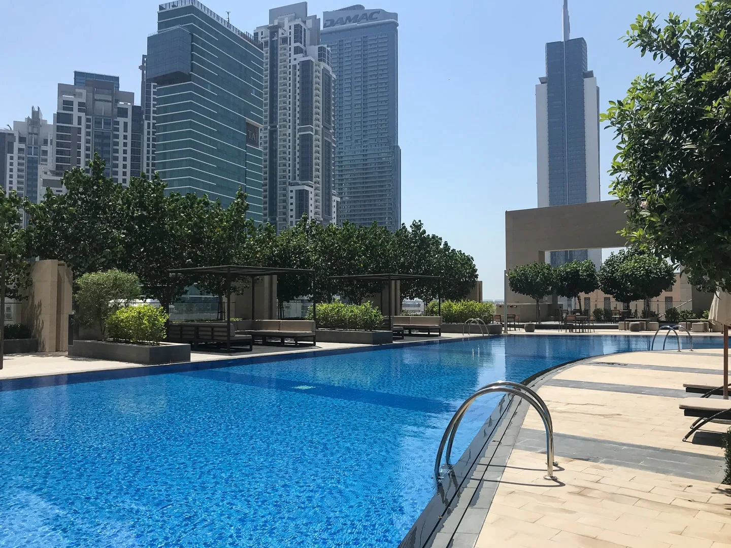 Una piscina al aire libre está junto a asientos con sombra, con edificios altos al fondo.