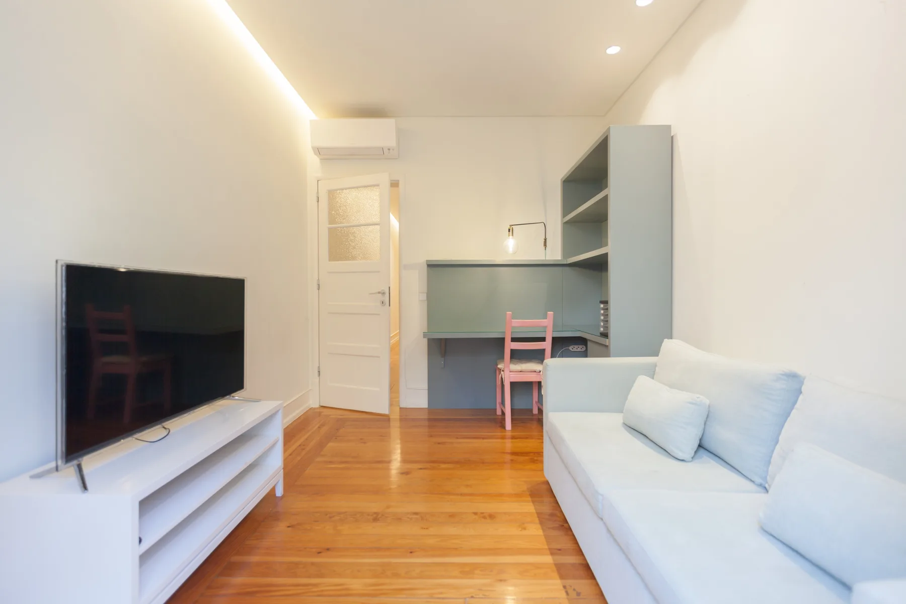 Una sala luminosa con un televisor, asientos claros y suelo de madera.