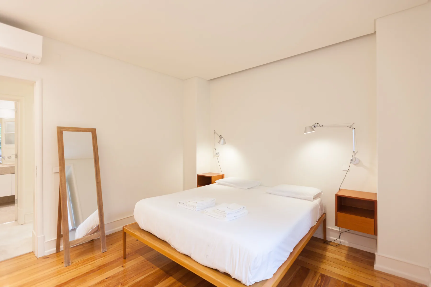 Dormitorio luminoso con cama, mesitas de noche y un espejo apoyado.