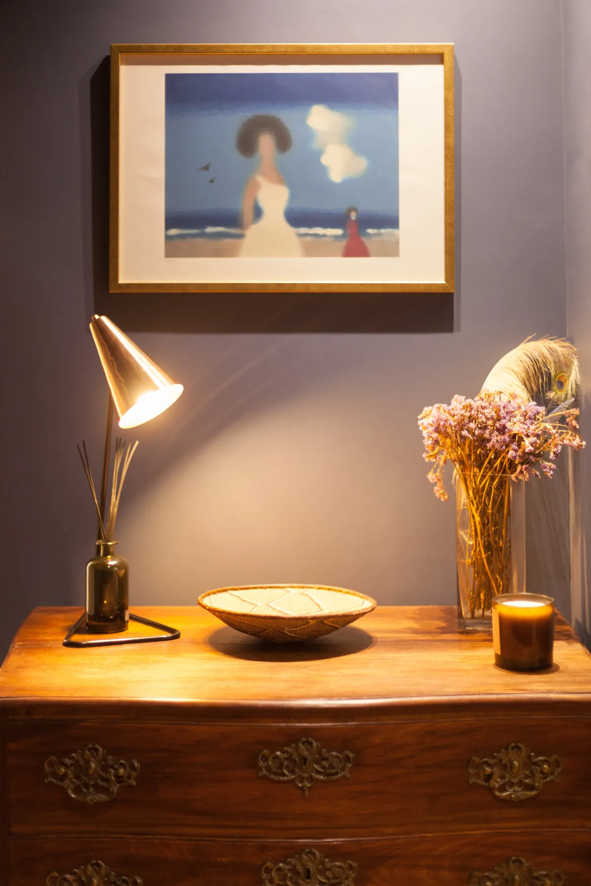 Una cómoda de madera sostiene una lámpara, un cuenco bajo, flores secas y una vela bajo un cuadro.