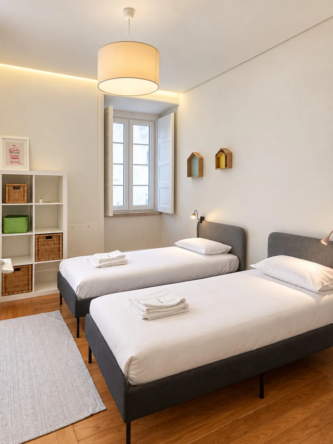 Un dormitorio luminoso con dos camas, una ventana y estanterías abiertas.