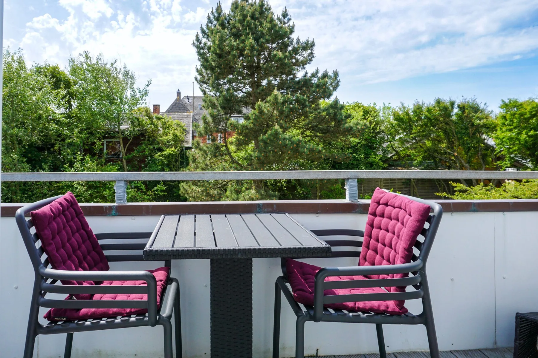 A balcony seating area with two chairs and a table overlooks leafy trees.