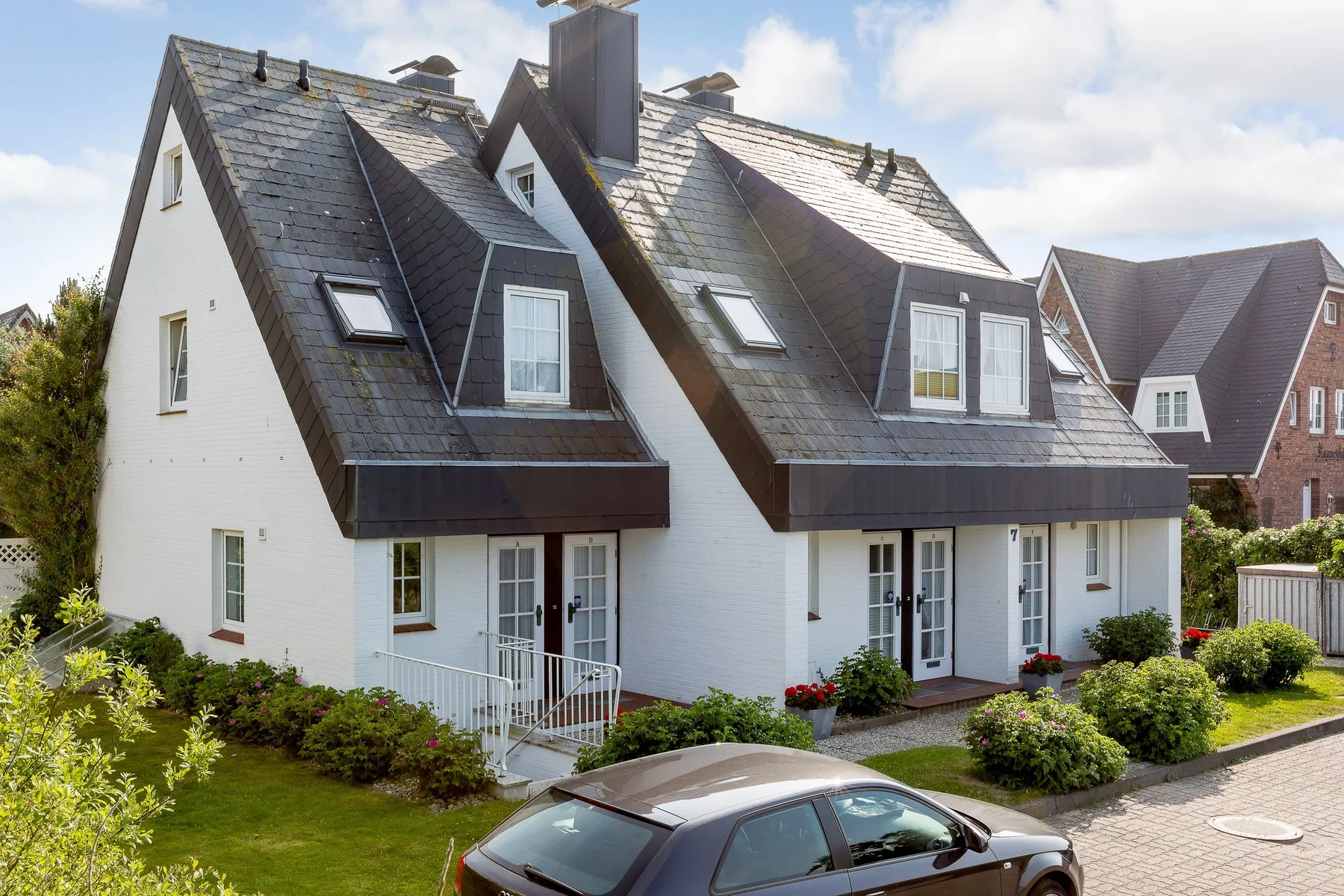 White houses with steep dark roofs stand beside a landscaped garden and paved drive.