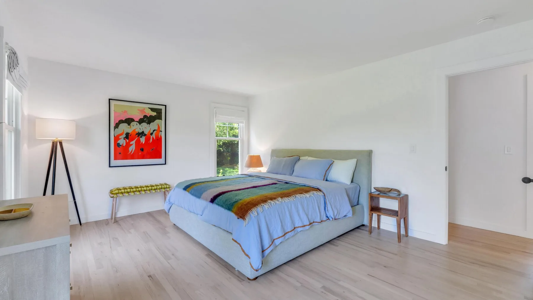 Un dormitorio luminoso con una cama, arte colorido y piso de madera clara.