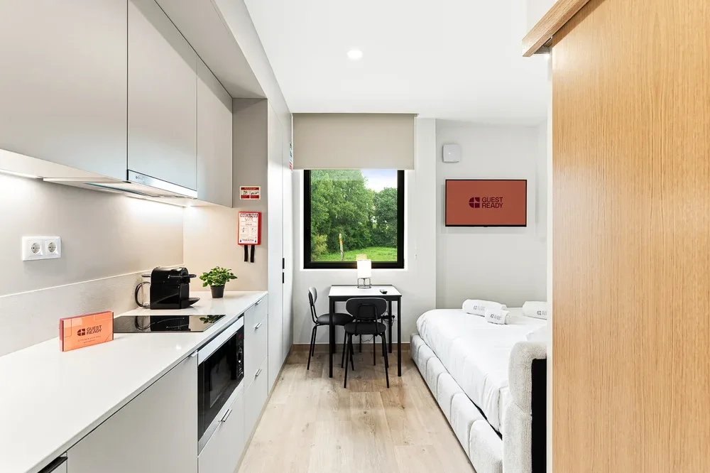 A bright room combines a kitchenette, dining table, and beds beside a large window.