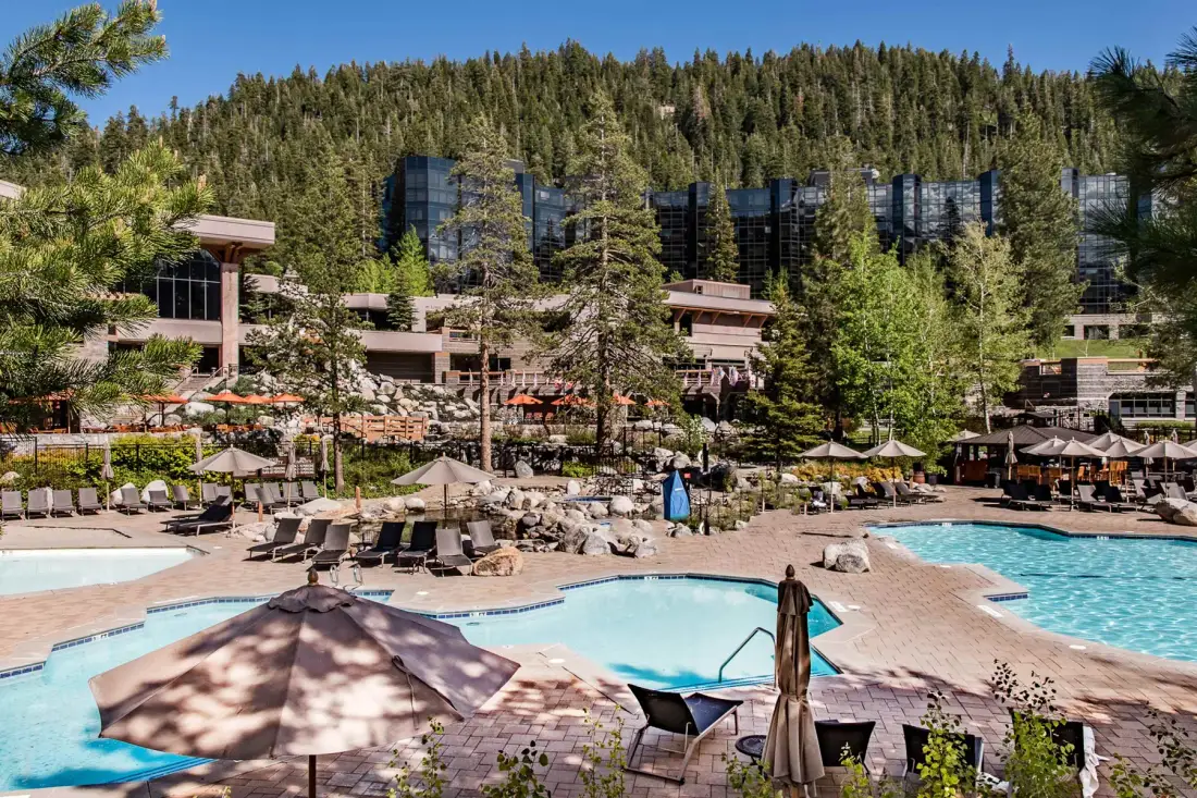 An outdoor pool area with lounge seating, umbrellas, and buildings against a wooded hillside.