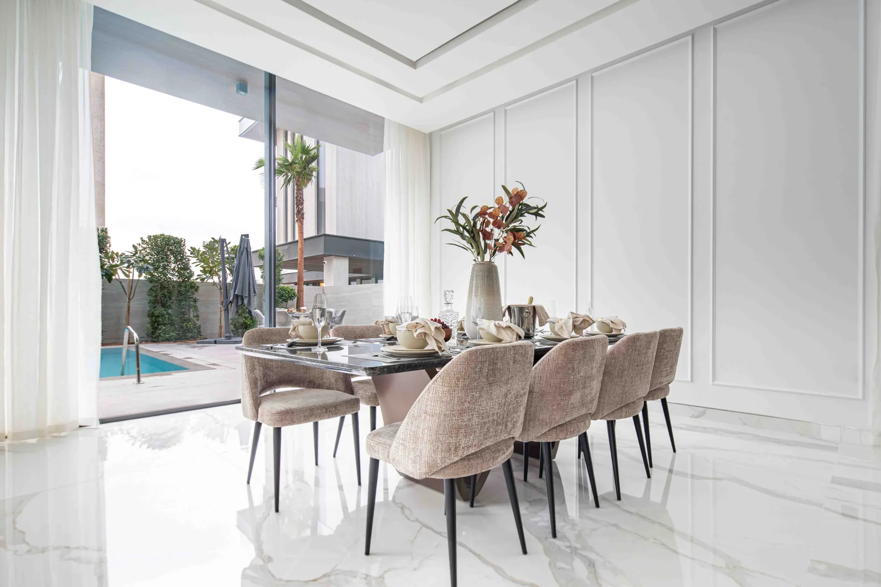 Un comedor luminoso con la mesa puesta junto a grandes ventanales con vistas a una piscina.