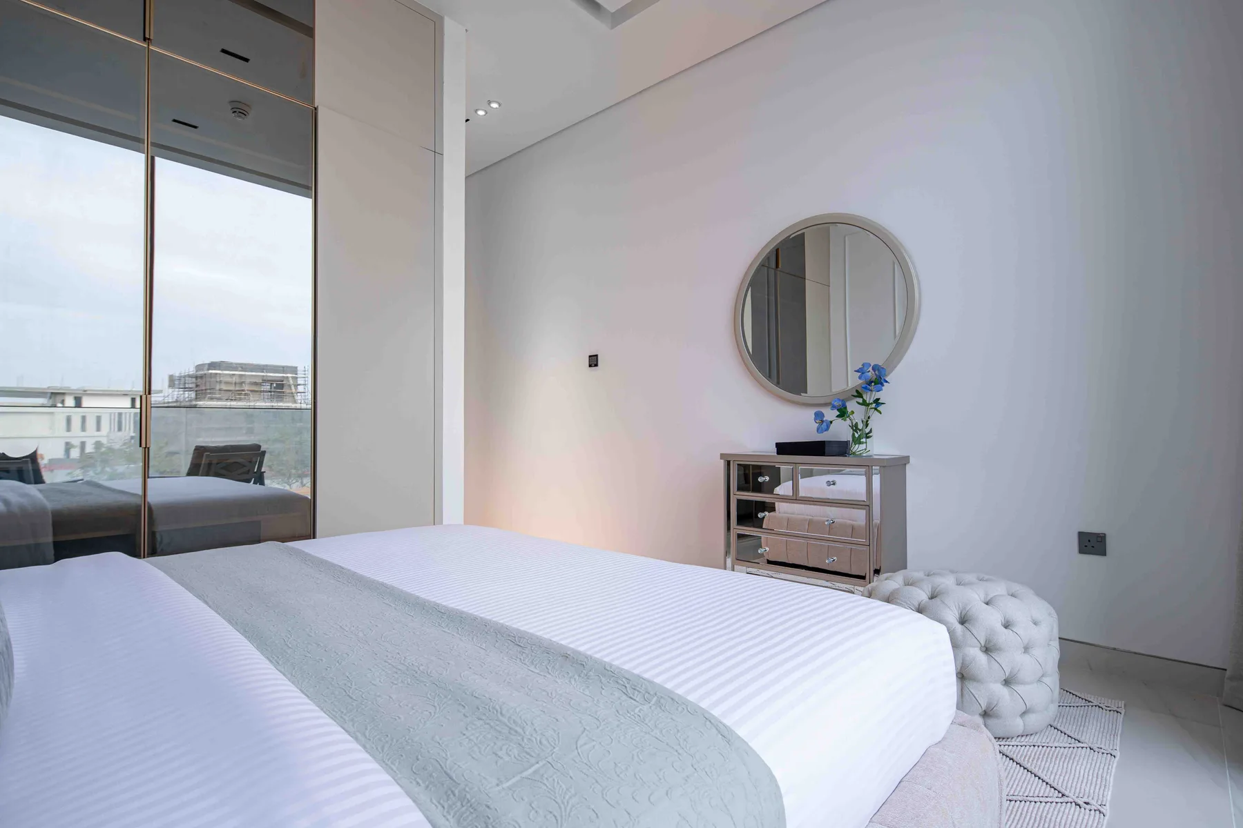 Un dormitorio luminoso con una cama bien hecha y un espejo redondo sobre una cómoda.