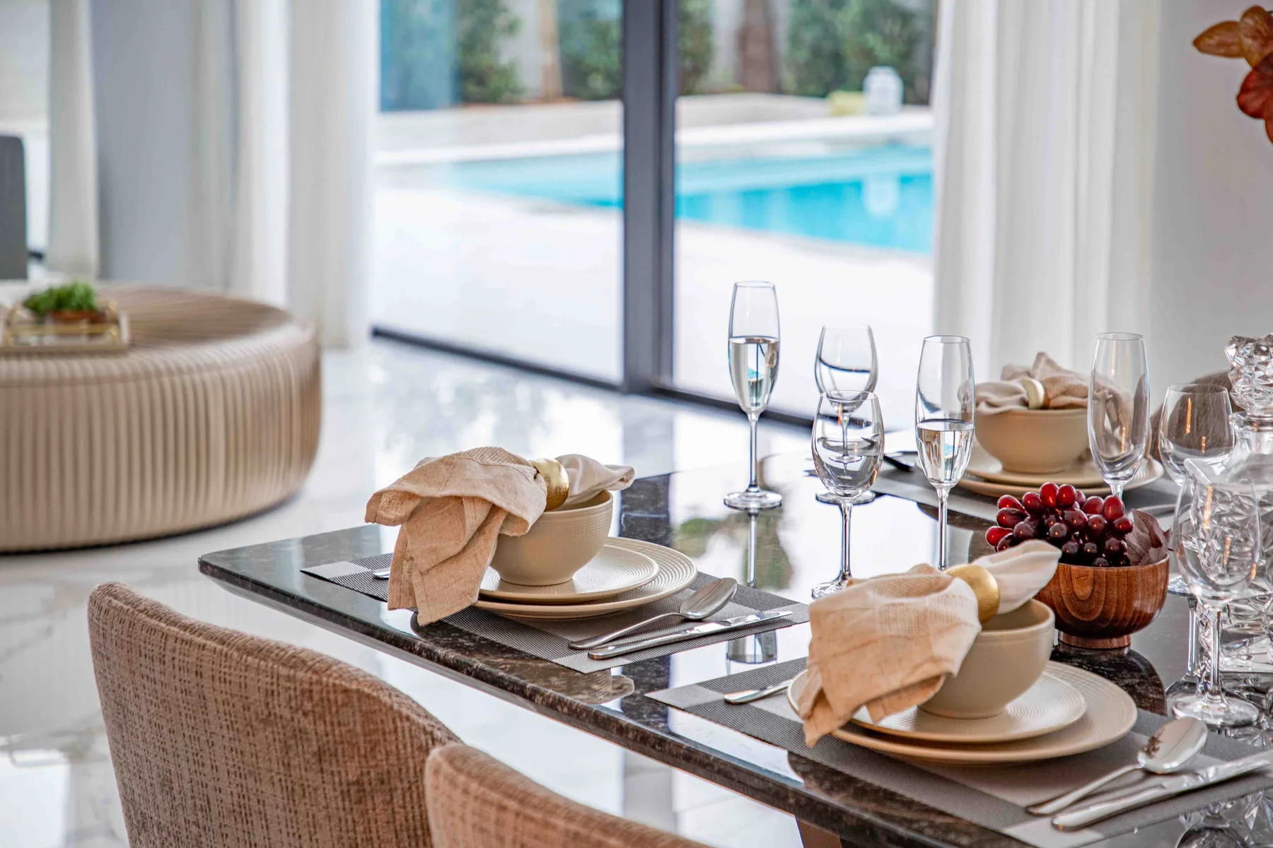 Una mesa de comedor está preparada con platos y copas junto a una ventana luminosa que da a la piscina.
