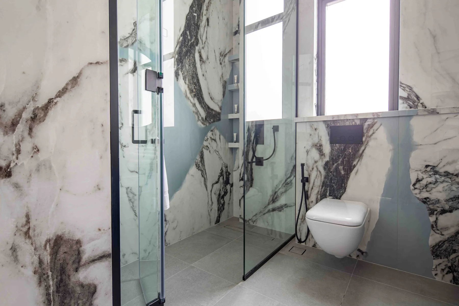 Un baño moderno con ducha de vidrio, inodoro suspendido y una ventana grande.
