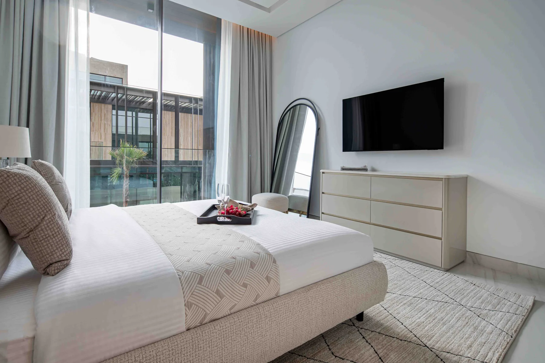 Un dormitorio luminoso con una cama, un televisor en la pared y ventanales.