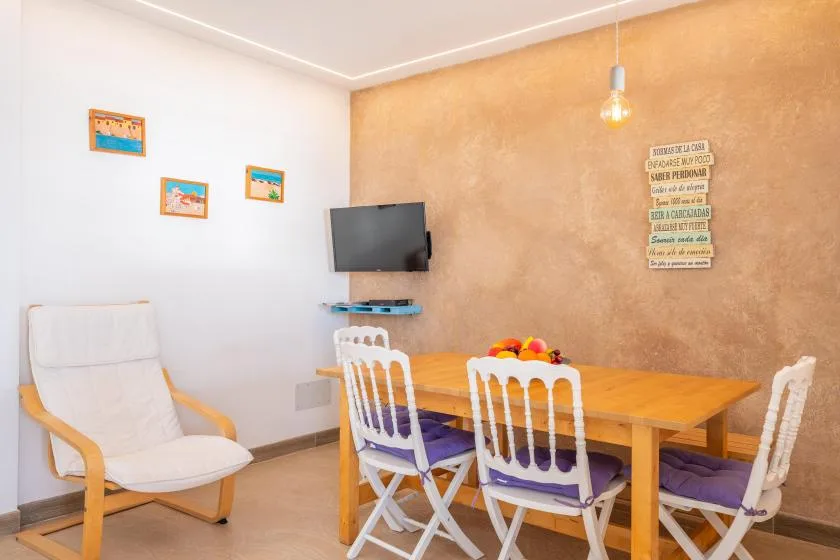 Un comedor con una mesa de madera, sillas blancas y un televisor montado en la pared.