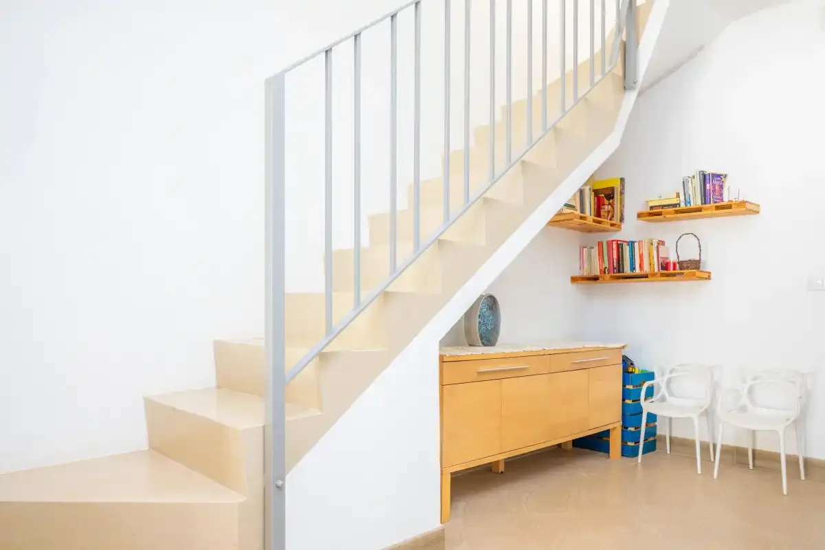 Un interior luminoso con una escalera, un mueble de madera y estantes en la pared.