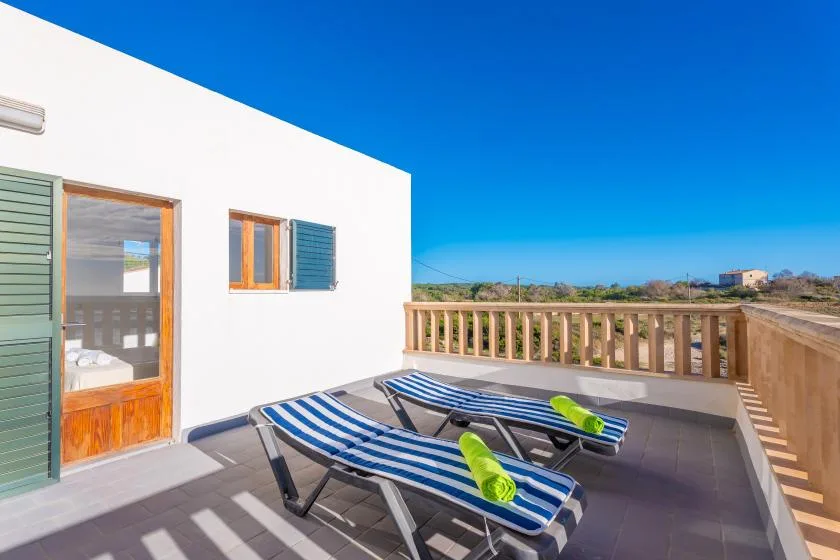 Una terraza soleada junto a un edificio blanco tiene tumbonas a rayas y una barandilla de madera.
