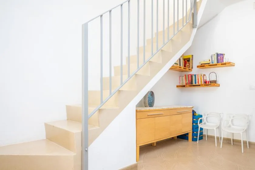 Un interior luminoso muestra una escalera, un mueble de madera, estantes y asientos blancos.
