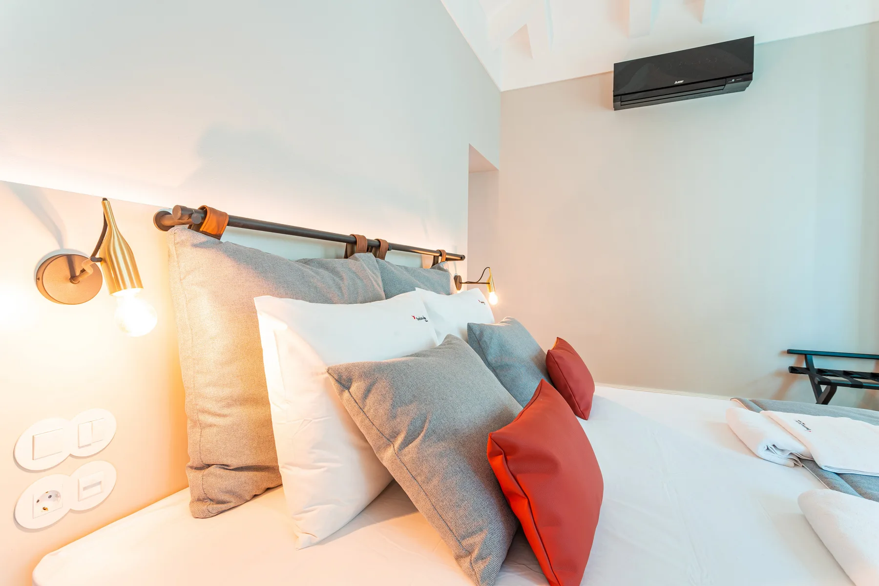 Un dormitorio luminoso con una cama vestida con ropa blanca y cojines grises y rojos.
