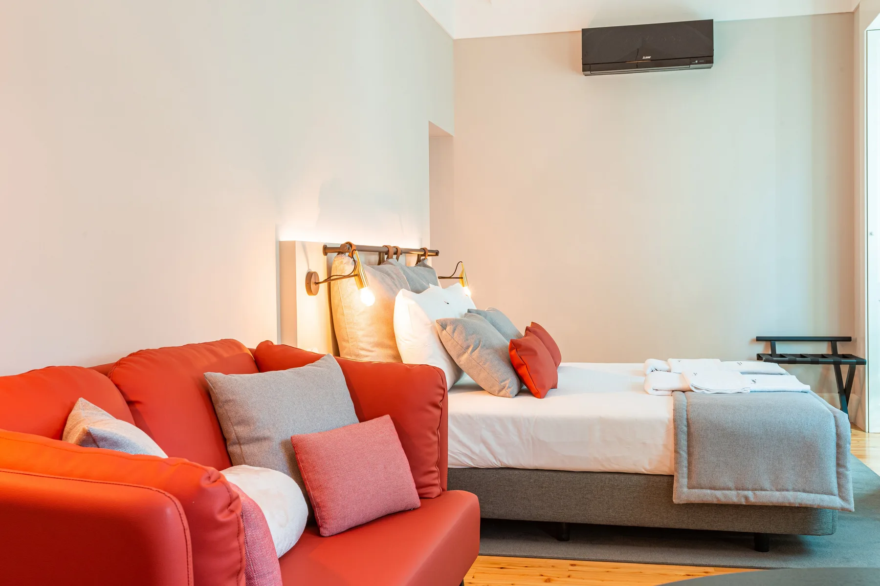 Zona de descanso de un estudio con una cama junto a un sofá rojo y un aire acondicionado en la pared.