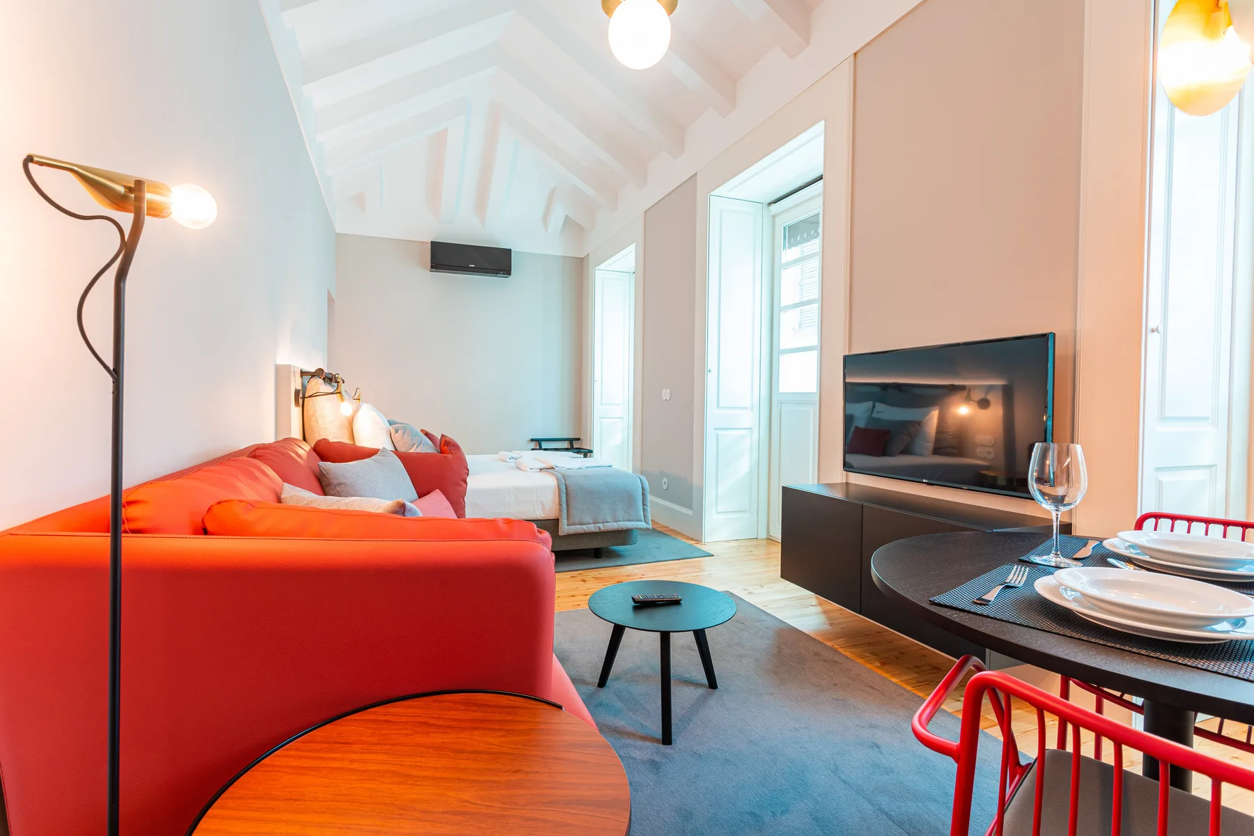 A bright studio living area has red seating, a television, and a dining table.