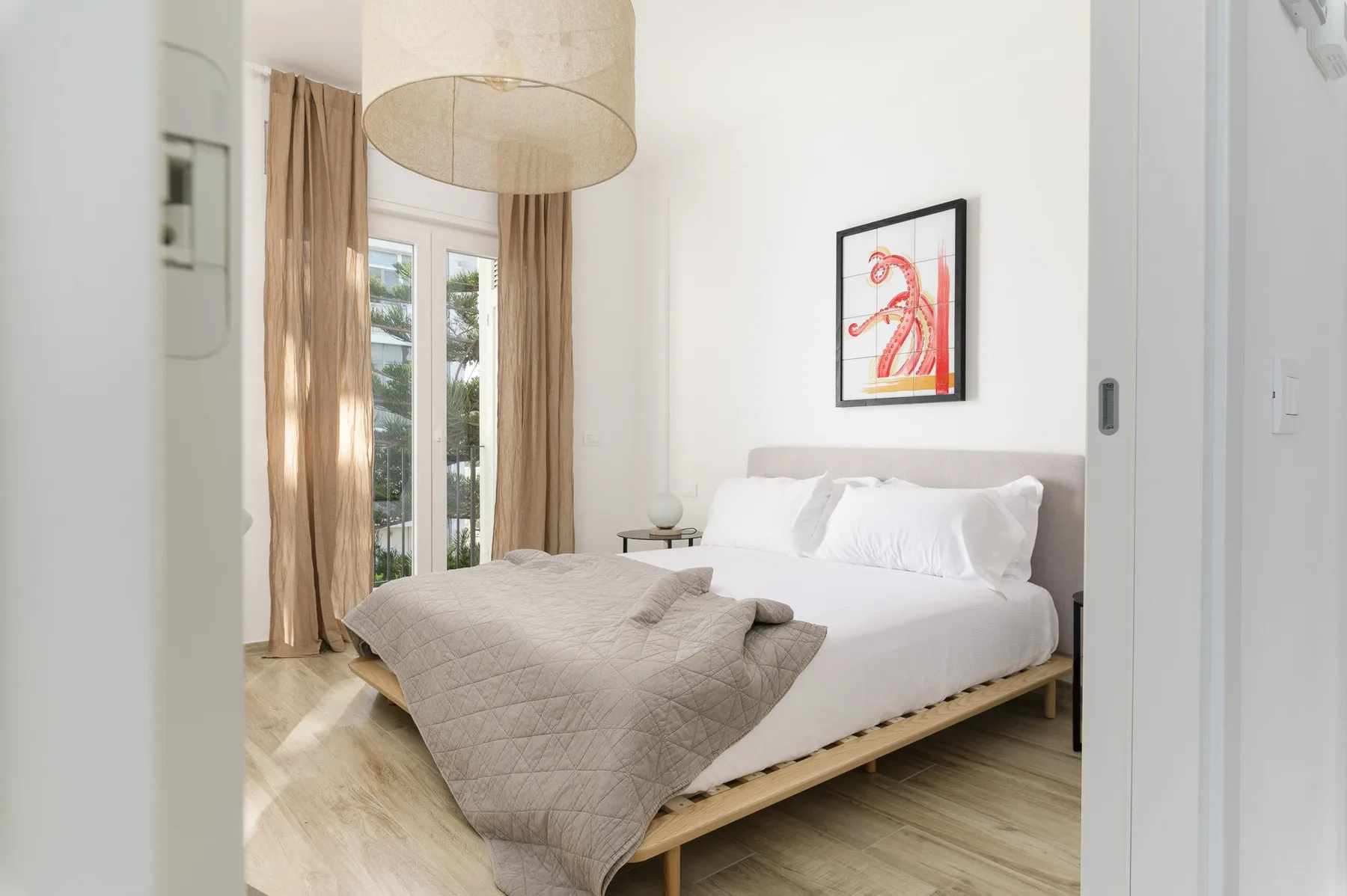 Un dormitorio luminoso con una cama bien tendida y ventanas altas con cortinas.