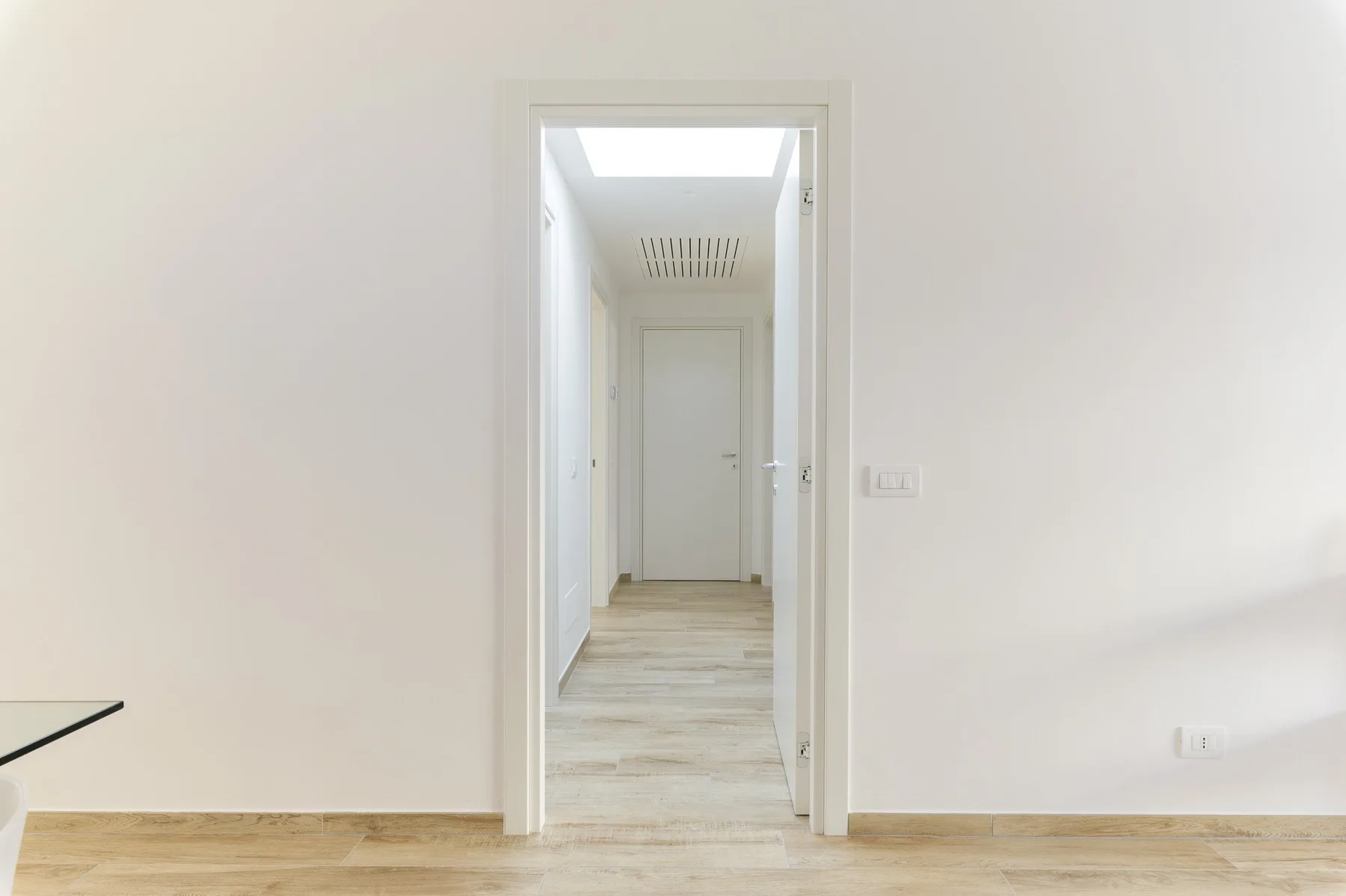 Un pasillo interior luminoso con suelo de madera clara y una puerta cerrada al fondo.
