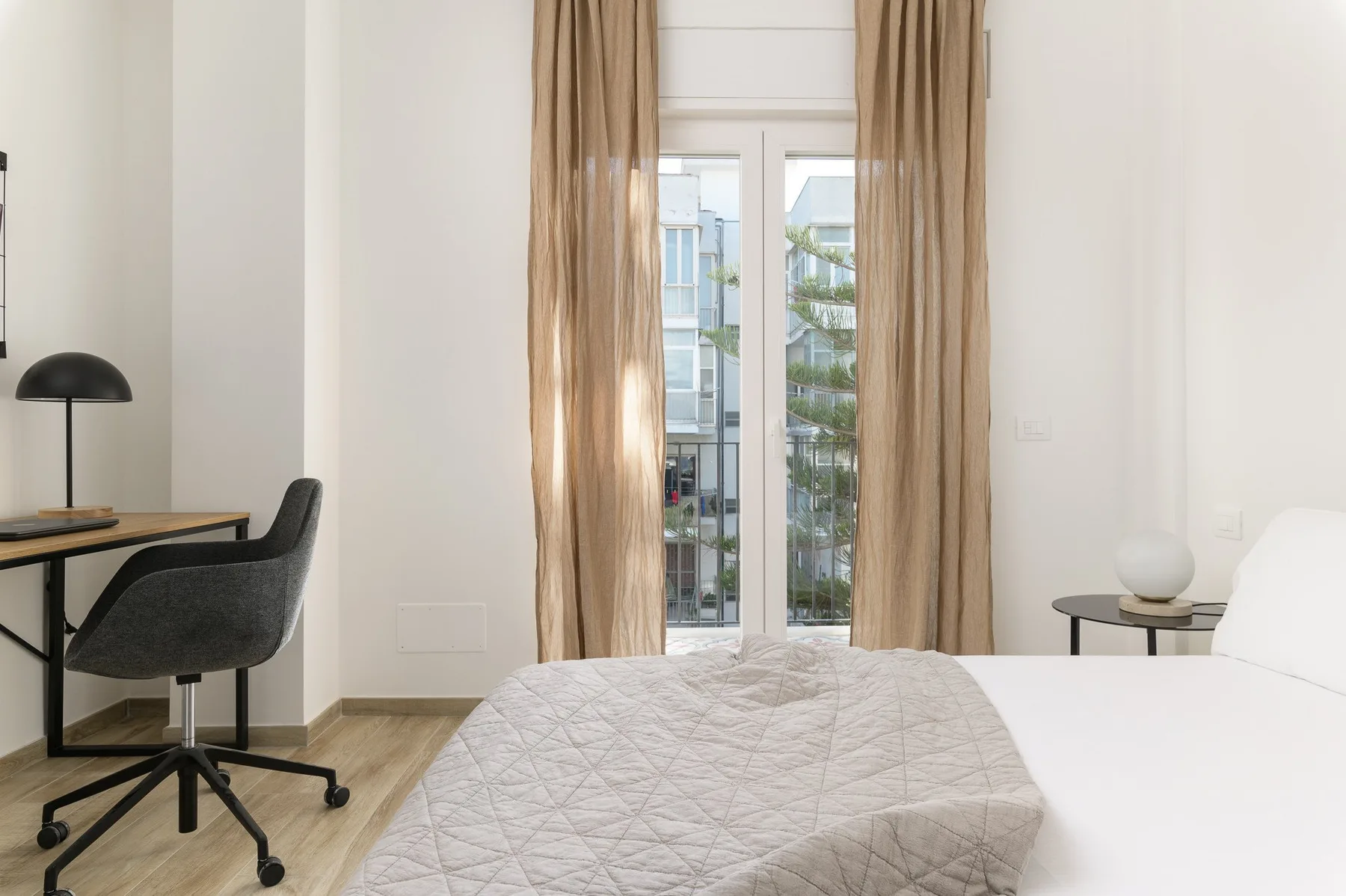 Un dormitorio con una cama junto a un escritorio y ventanas altas con cortinas.