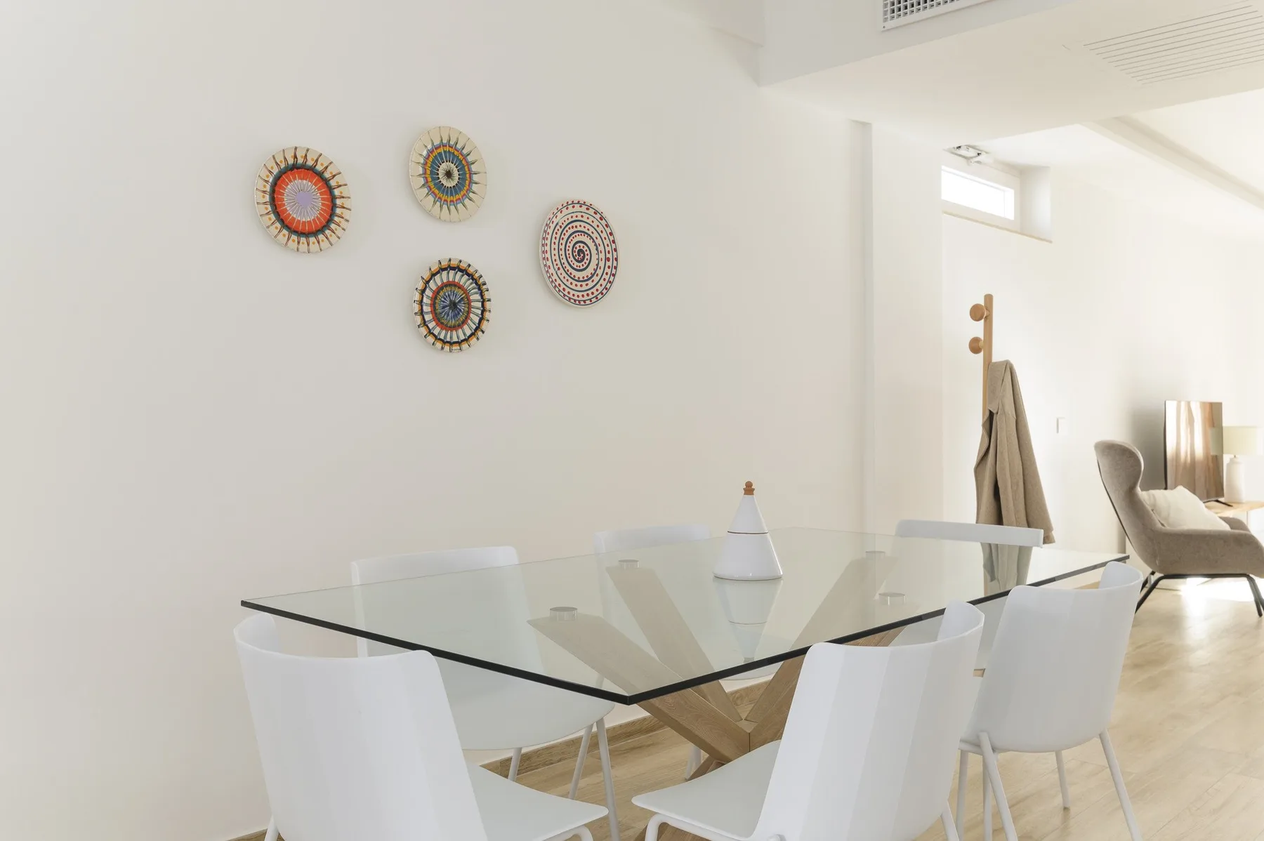 Un comedor luminoso con una mesa de vidrio, asientos blancos y adornos coloridos en la pared.