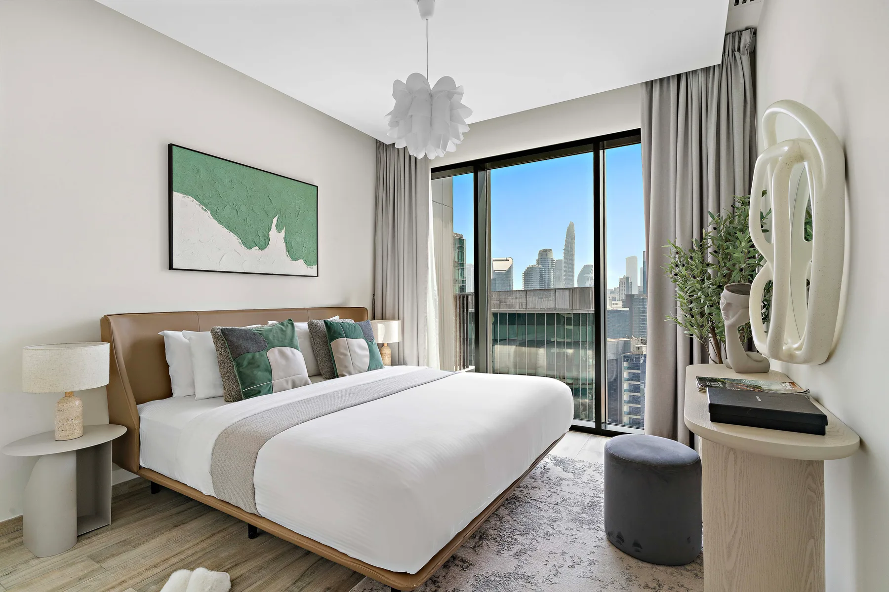 A bright bedroom with a neatly made bed beside large windows overlooking the city.