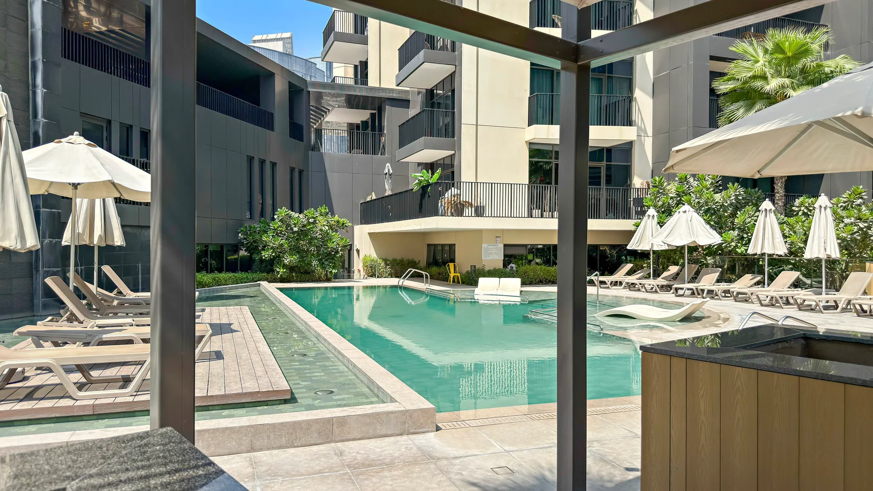 An outdoor swimming pool sits among modern buildings and rows of lounge seating.