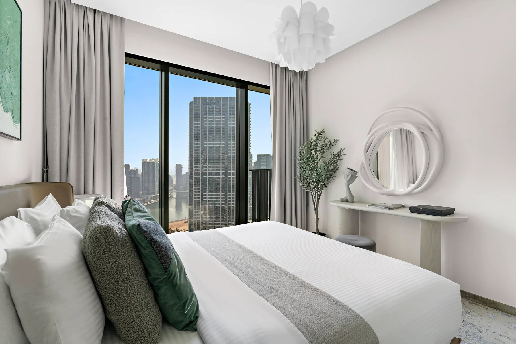 A bright bedroom with a bed beside large windows overlooking city buildings.