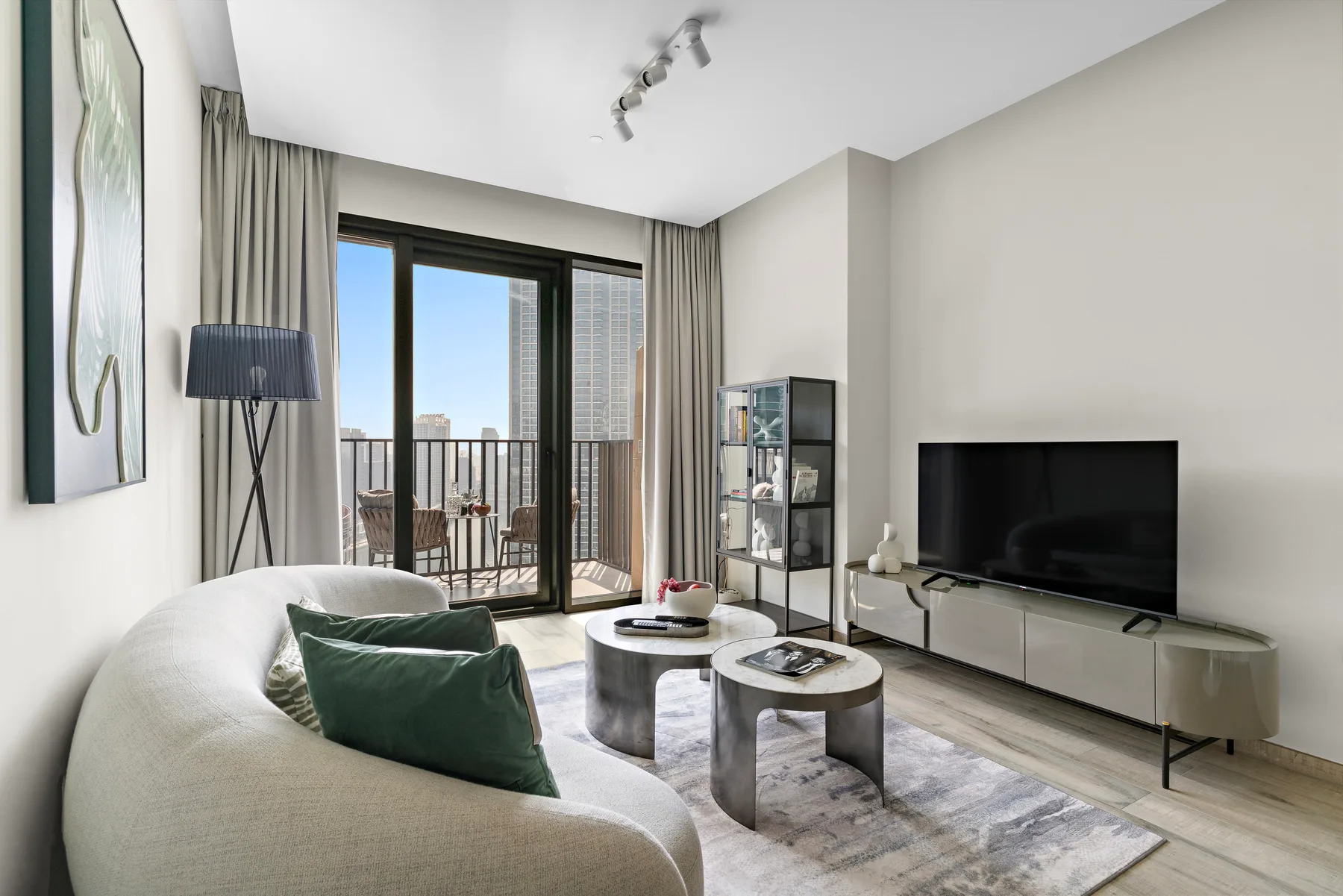 A bright living room with seating, coffee tables, and a television beside balcony doors.