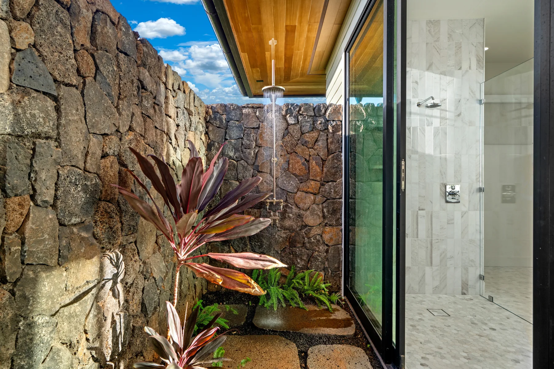 An outdoor shower beside a stone wall and leafy plants.