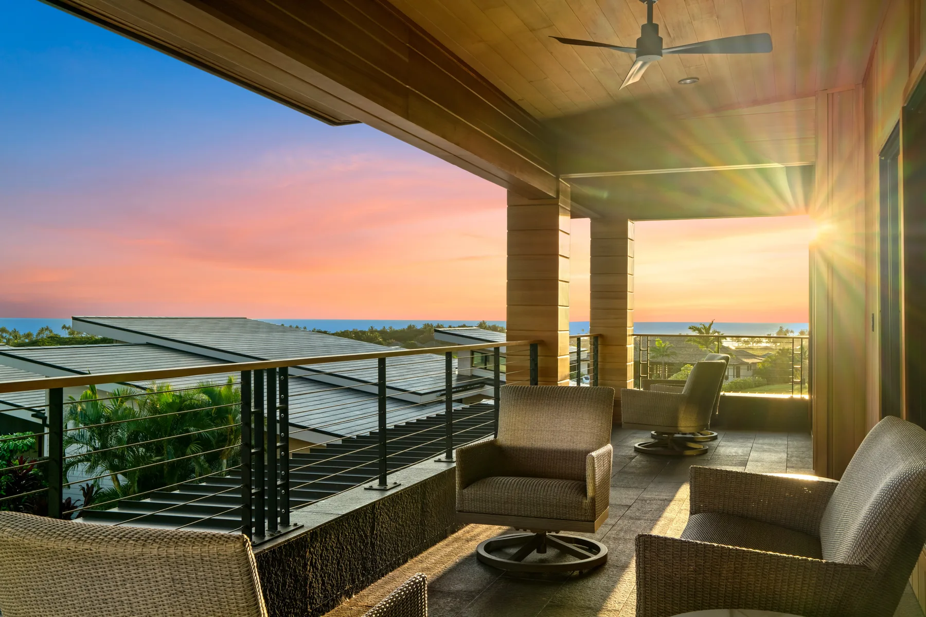 A covered balcony with outdoor seating overlooks rooftops and the ocean at sunset.