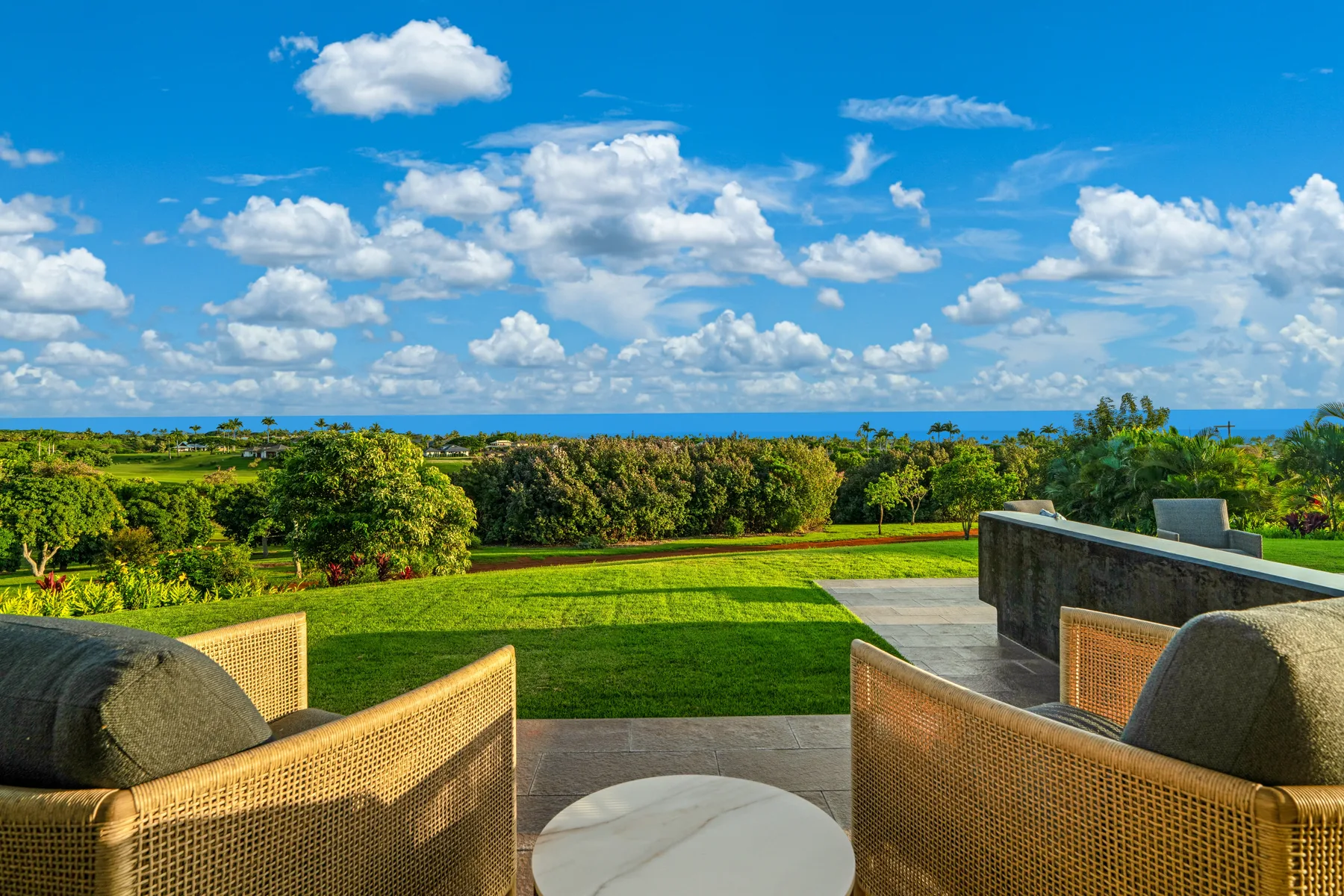 A patio seating area overlooks a green lawn and the ocean beneath a blue sky.