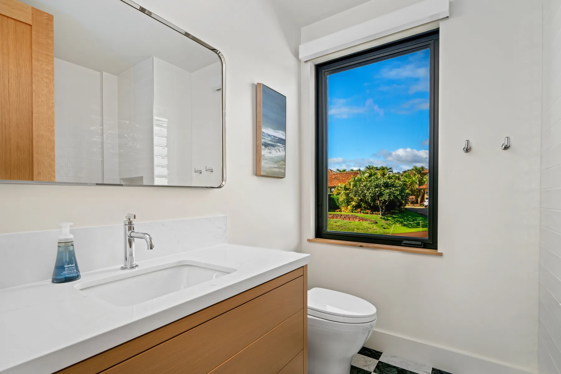 A bright bathroom with a vanity, toilet, and a large window overlooking greenery.
