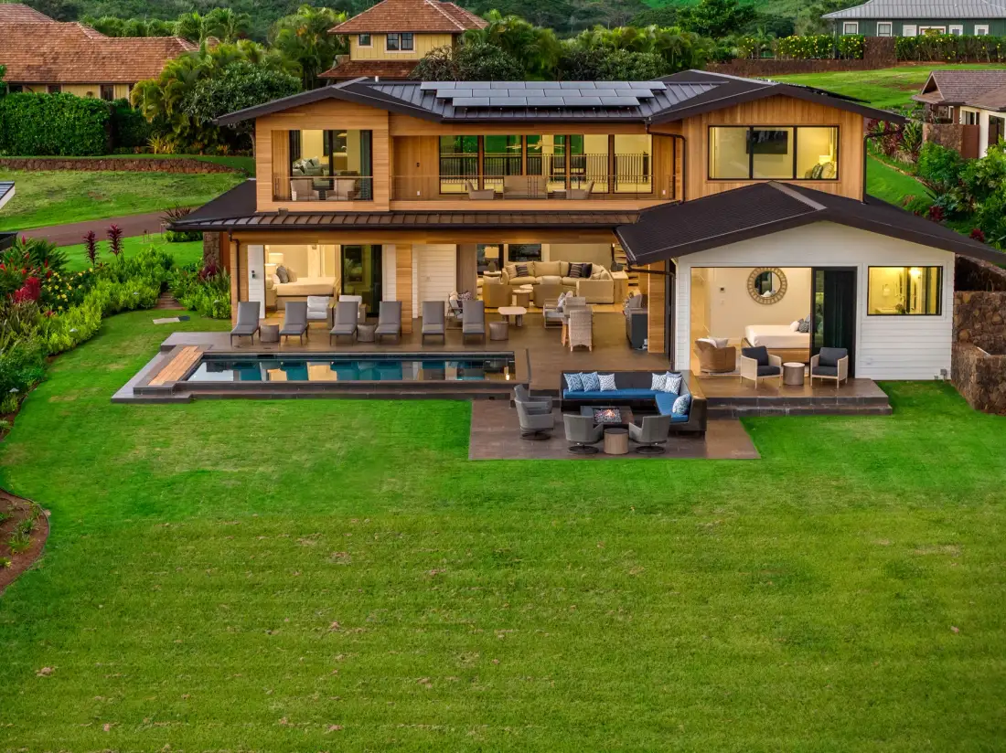 A large modern house overlooks a green lawn, with a swimming pool and patio seating.