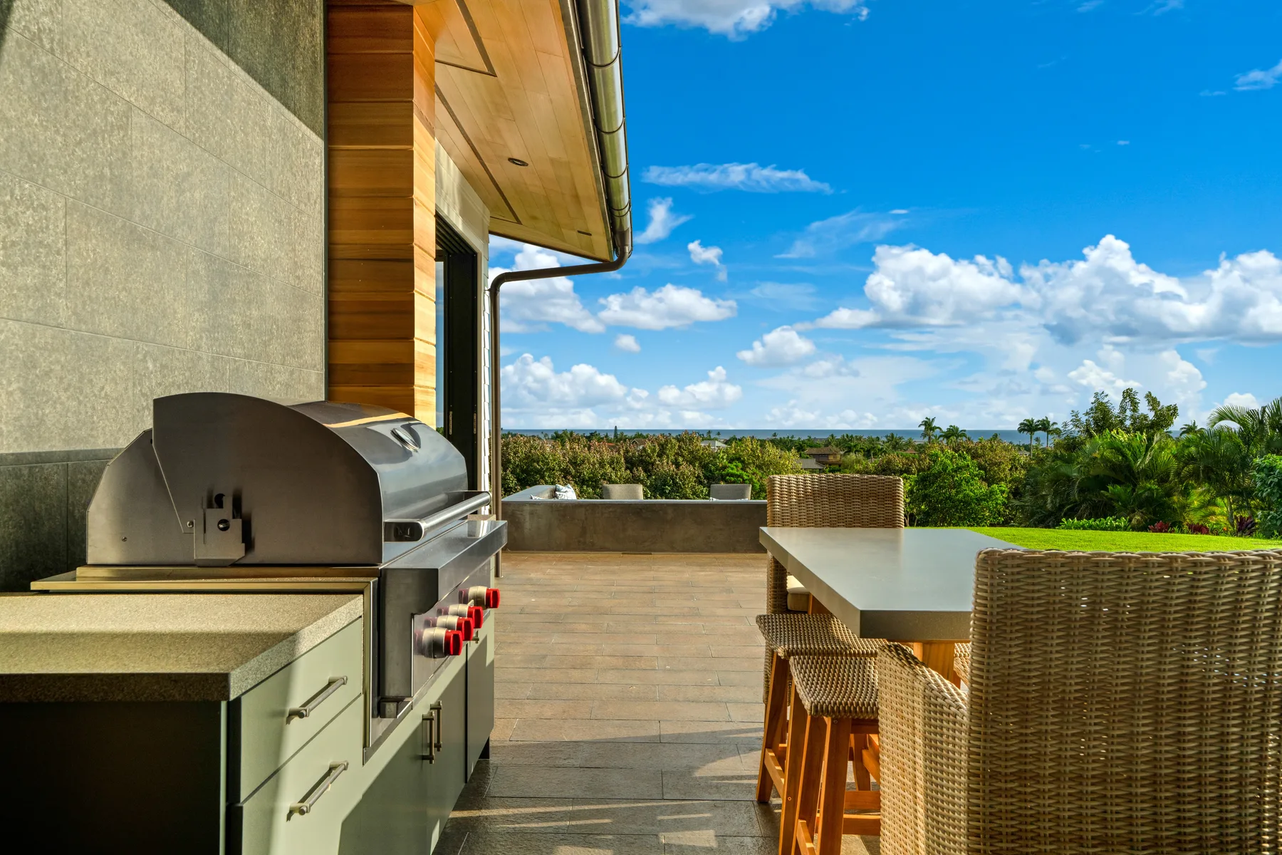 An outdoor dining patio with a built-in grill and woven seating overlooks greenery.