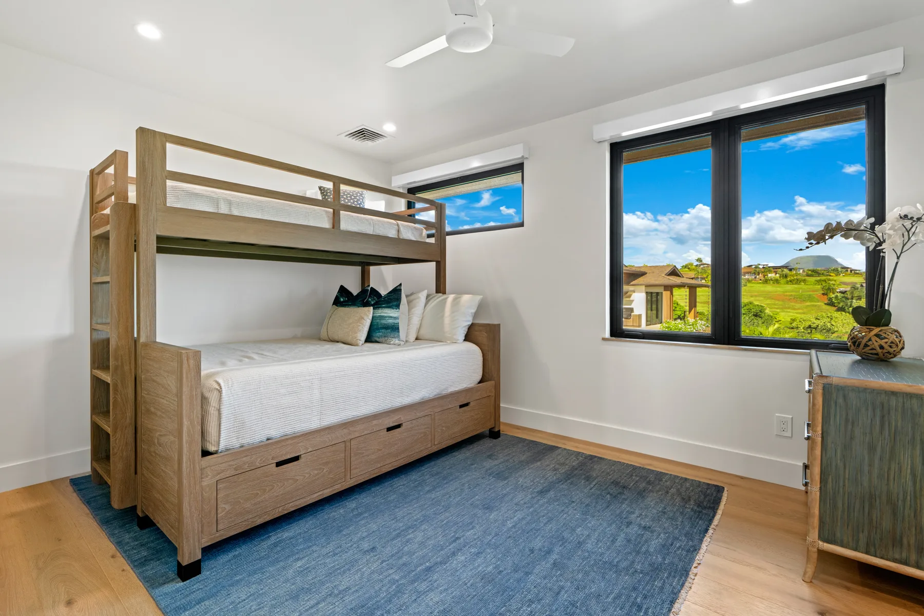A bedroom with a wooden bunk bed, blue rug, and windows overlooking greenery.