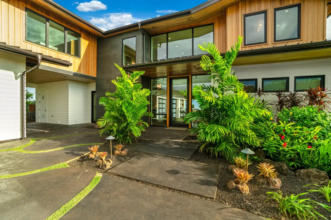 A modern home exterior with a paved entrance and lush tropical landscaping.